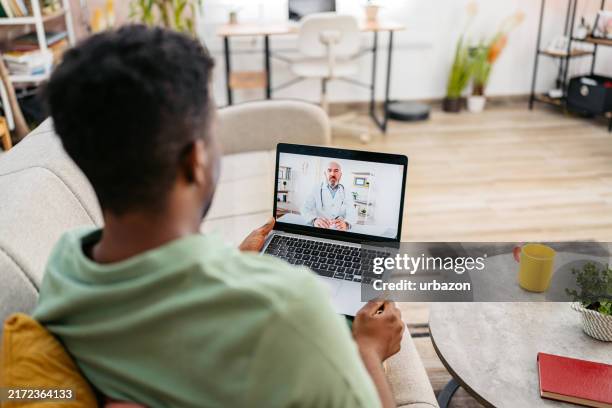 young man having a doctor's appointment on the laptop on the sofa - telemedicine stock pictures, royalty-free photos & images