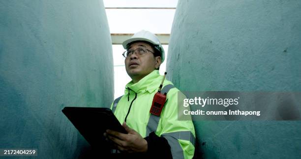 the chief industrial engineer wearing a reflective green safety jacket is using a digital tablet and walkie-talkie to communicate with his team. they are rechecking the liquid flow system in huge water tanks at the side of the factory. - storage tank stock pictures, royalty-free photos & images