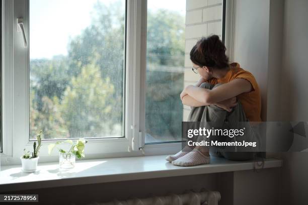 sad upset teenager sitting on windowsill hugging knees looking out window, lonely teen girl at home. adolescent loneliness - verlaten stockfoto's en -beelden