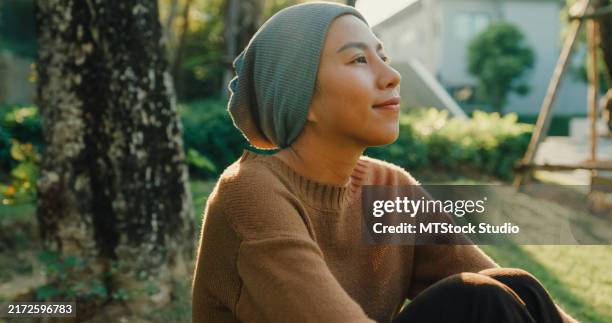 nahaufnahme einer jungen asiatischen frau, die an krebs erkrankt ist, sitzt ruhig auf dem gras, in gedanken versunken und genießt die ruhe eines üppigen gartens, der ein gefühl von frieden und genesung widerspiegelt. leben mit krebs. - überleben stock-fotos und bilder