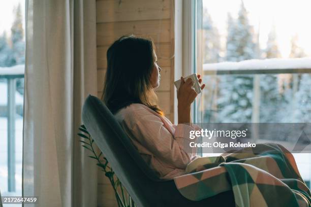 alone with thoughts in armchair of winter house. woman sips coffee and looks out window at snowy sunrise - schaukelstuhl stock-fotos und bilder