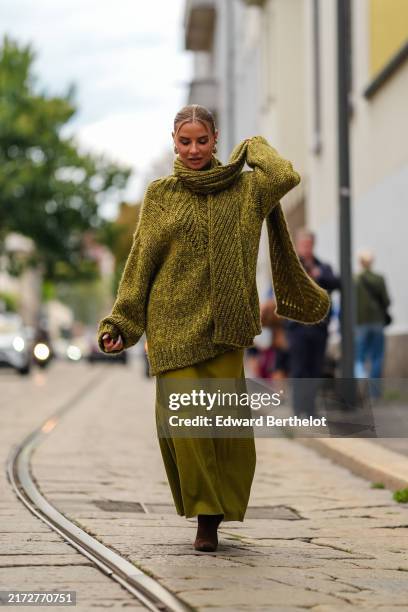Veronica Ferraro wears gold earrings, dark green cotton knit oversized sweater, dark green cotton knit, dark green cotton knit oversized scarf, dark...