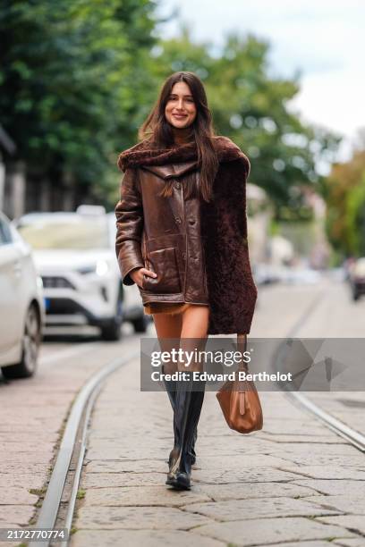 Belen Hostalet wears dark brown oversized scarf, shiny dark brown oversized leather jacket, light brown leather bag, shiny black leather boots,...