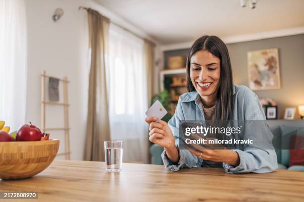 a young, cheerful woman using her smartphone to shop online and holding a credit card. - credit card stock pictures, royalty-free photos & images