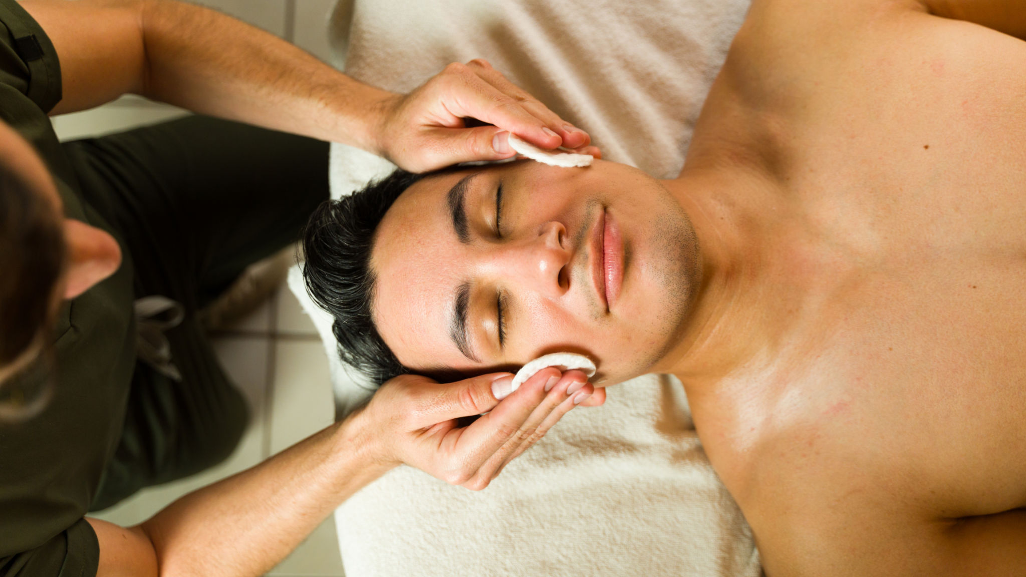 Male beautician is giving a facial treatment to a man lying on a massage table in a spa Male beautician is giving a facial treatment to a man lying on a massage table in a spa