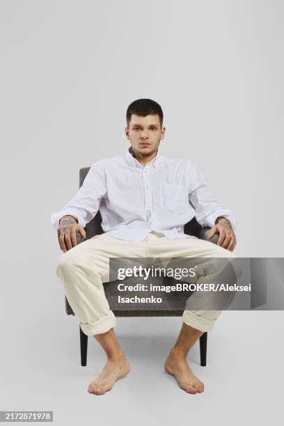 full length studio portrait of young barefoot man in white shirt and ivory trousers sitting in armchair - white pants stock pictures, royalty-free photos & images