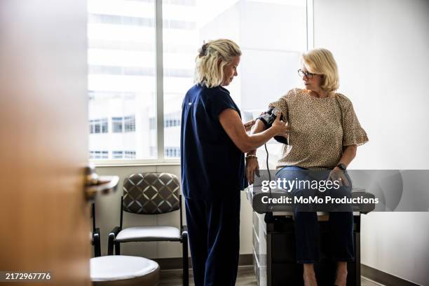 female nurse in scrubs measuring senior woman's blood pressure in exam room - bluthochdruck stock-fotos und bilder