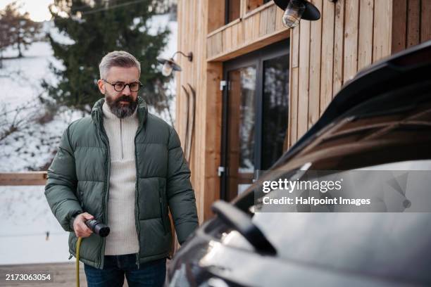 man charging electric car in front his cabin in mountains. electric vehicle with charger in charging port. man driving electric car on winter holiday in mountains. - ice charger imagens e fotografias de stock