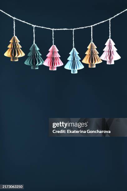 colorful paper tree ornaments hanging on a string. minimalist holiday décor with multiple colors of christmas trees on a dark background and copy space. - december stockfoto's en -beelden
