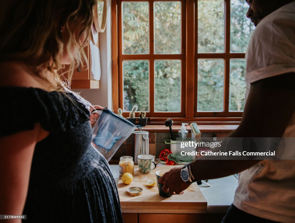 A pregnant woman pours some water from a water filtration jug as she stands in a kitchen with her partner