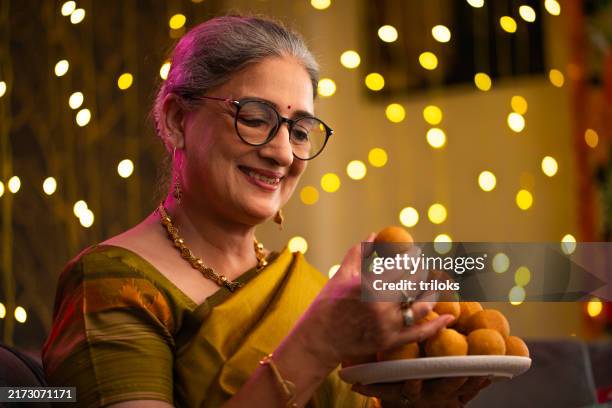 senior woman showing laddoo while celebrating diwali at home - eyewear stock pictures, royalty-free photos & images