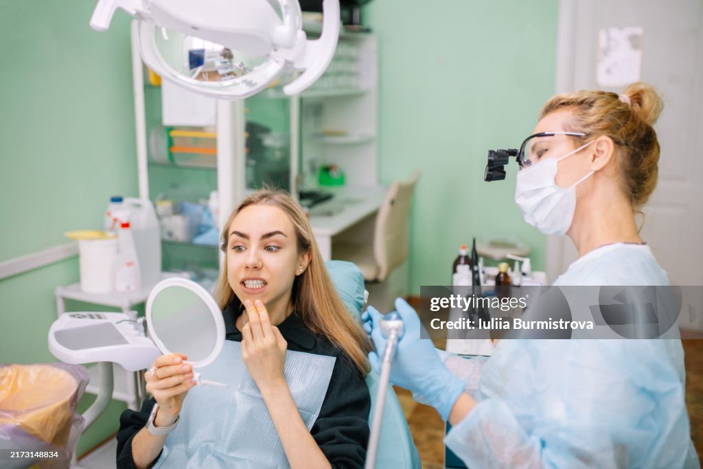 Charming young lady examines refreshed smile in handheld mirror while skilled dentist prepares for next phase of procedure