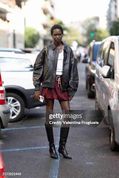 Guest wears a white tank top, a red burgundy mini puffy skirt, black leather boots and a brown leather jacket outside Max Mara show during the Milan...