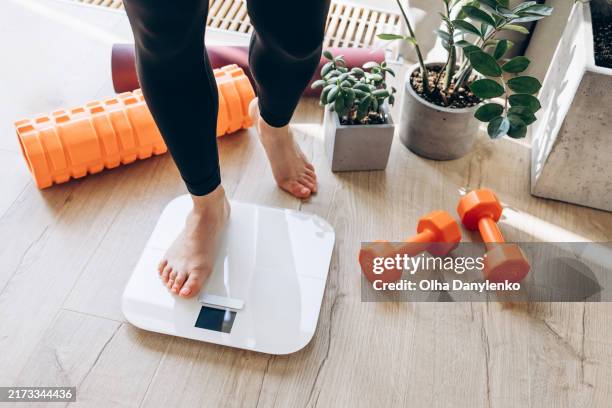 woman steps on a weighing scale with dumbbells and fitness equipment in the background. healthy lifestyle concept - perte photos et images de collection