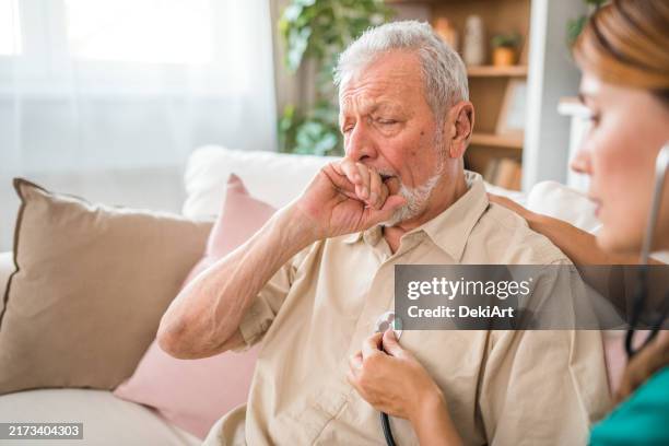 ein älterer mann hustet, als eine krankenschwester während eines hausbesuchs seine lunge mit einem stethoskop untersucht - herzton abhören stock-fotos und bilder