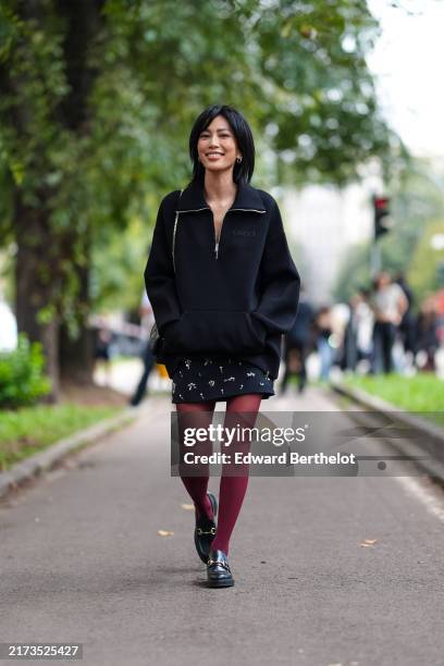Molly Chiang wears black oversized sweater, black mini dress with silver detail, dark red sheer mesh tights, shiny leather loafer moccasin leather...