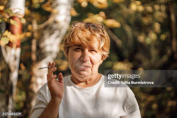blond-haired senior lady with warm smile pauses for smoke in tranquil ambiance of sunlit woods - squinting stock pictures, royalty-free photos & images