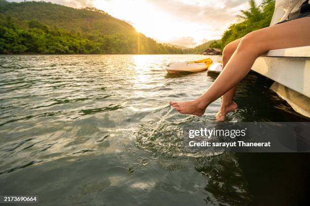 woman relaxes by the lake sitting on the edge of a wooden jetty, swing feet near the water surface - ponton photos et images de collection