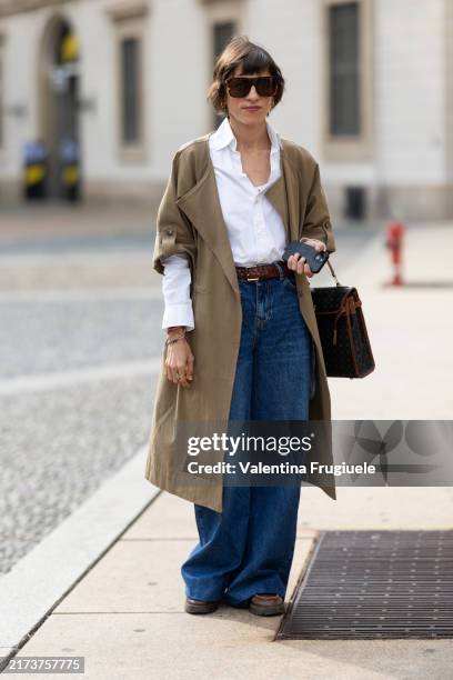 Guest wears brown sunglasses, brown leather loafers, a brown leather belt, a white shirt, a beige trench, blue wide leg jeans and a black and brown...