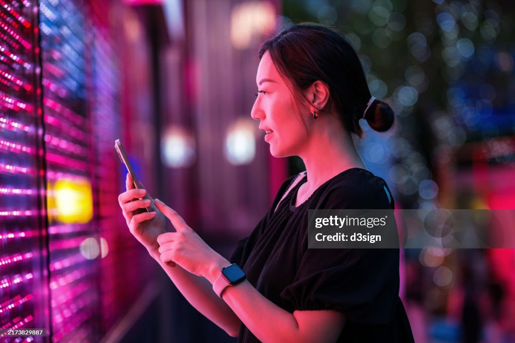 Confident young Asian businesswoman using smartphone, standing against illuminated digital display on the street in city at night. Concept of VR and AR technologies. Metaverse and futuristic data. People, AI, innovation and technology