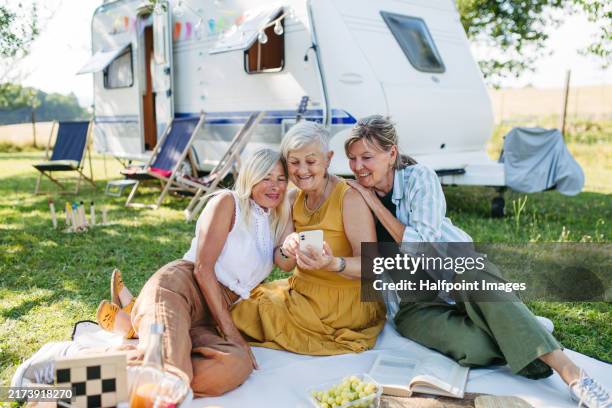 senior best friends at camping trip in nature, sitting on picnic blanket and taking selfie. adventurous older women going on road trip with caravan, enjoying peaceful moment. - caravan stockfoto's en -beelden