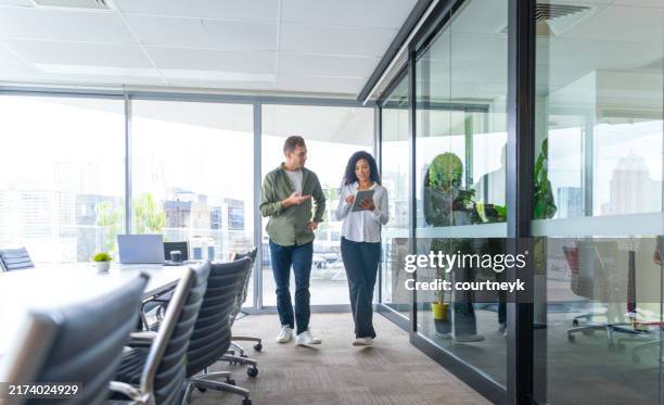 man and woman walking and talking in the office brainstorming or discussing a project. they are in a conference room holding a digital tablet - human resources stock pictures, royalty-free photos & images