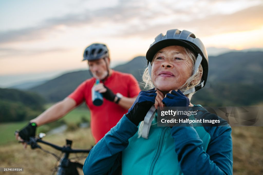 Portrait of beautiful senior couple on bike ride in autumn nature. Taking break, drinking water from sport bottle and fastening helmet.