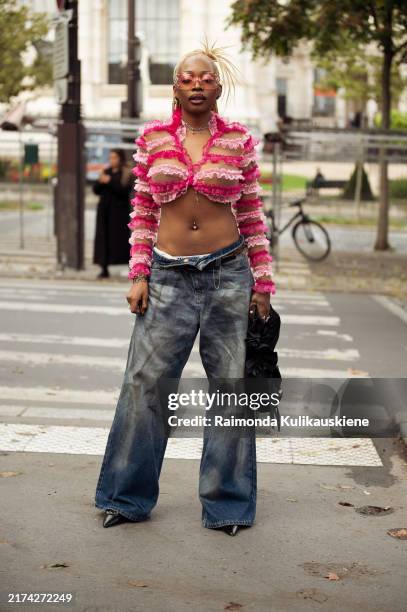 Guest wears wide blue jeans, pink and transparent fabric ruffled long sleeve cropped top, pink glasses and black bag outside the Anrealage show...