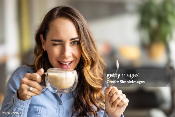 a cute young woman holds a mug with cappuccino in the other hand a spoon and winks - parpadear fotografías e imágenes de stock