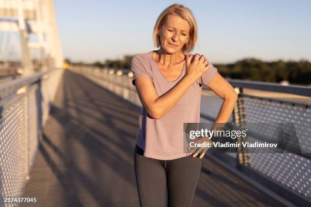 frau mit schmerzen hält schulter auf brücke während der goldenen stunde - schmerz stock-fotos und bilder