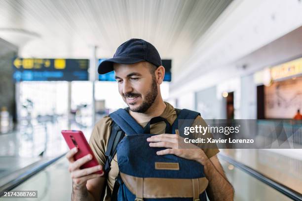 portrait of smiling young traveler man with backpack using smart mobile phone on moving walkway. - travel check in stock pictures, royalty-free photos & images
