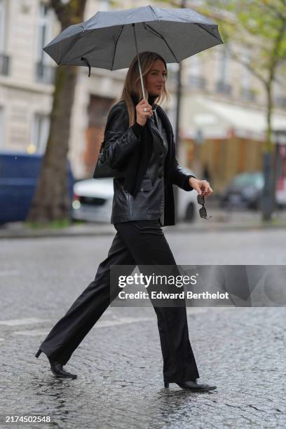 Guest wears shiny black oversized blazer jacket, black pants, black sunglasses, black pointed toe leather heels, outside Cecilie Bahnsen, during the...