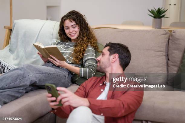 young woman reading a book while her boyfriend is using his smartphone on the floor - materiale cartaceo foto e immagini stock