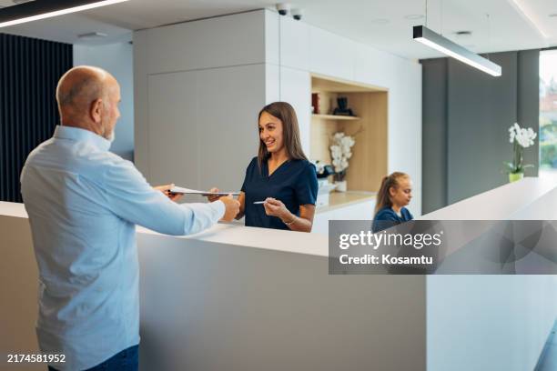 un hombre mayor entrega el formulario médico que llenó a la recepcionista de la clínica - clinica-medica fotografías e imágenes de stock