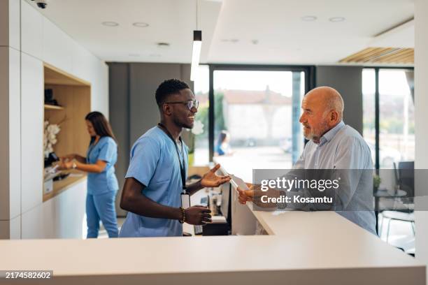 a smiling male receptionist in a modern clinic is talking to a male senior patient at the reception desk - reception bildbanksfoton och bilder