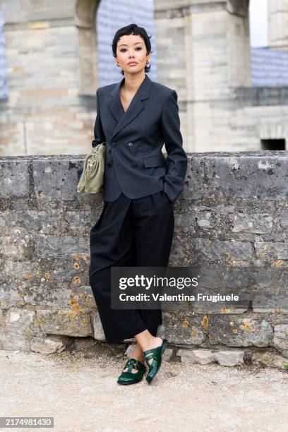 Guest wears gold hoop earrings, a light green leather pouch bag, green leather shoes, a black midi skirt and a grey blazer outside Loewe show during...