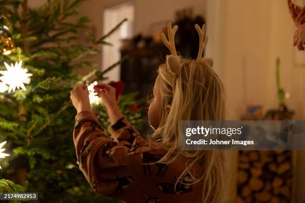 girl with reindeer antlers headband decorating star shaped lights on christmas tree at home - décembre photos et images de collection