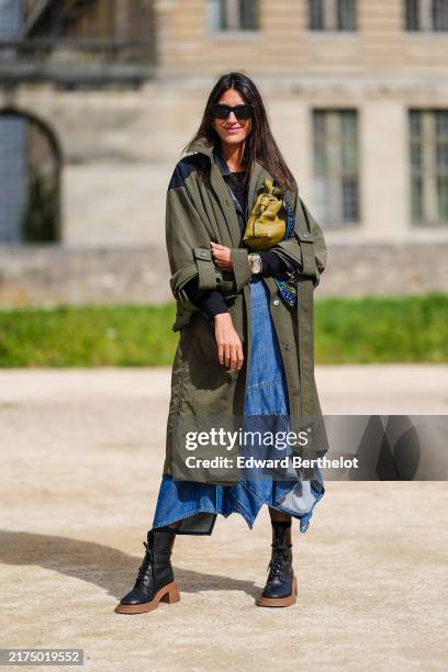Guest wears black sunglasses, dark green olive trench coat, light green leather bag, a silver watch, light blue denim jean flowy midi skirt, shiny...
