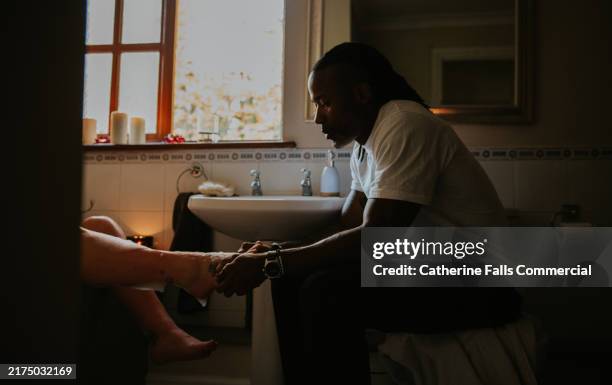 a man perches on a toilet seat in a domestic bathroom, giving his partner a foot rub as she dangles her feet out of the bath tub. - alternative therapy stock pictures, royalty-free photos & images