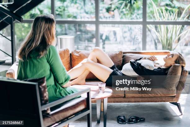 young woman is lying on couch with pillow, while another woman is sitting nearby. concept of therapy session, a conversation, or relaxation at home. - psychiatrists couch stock pictures, royalty-free photos & images