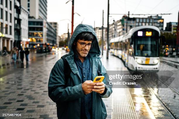turista adulto medio in una serata nuvolosa e piovosa utilizzando l'app per smartphone mentre aspetta il tram - provincia di anversa belgio foto e immagini stock