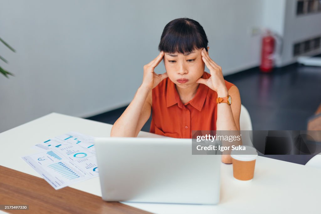 Stressed Asian Businesswoman Sitting at Desk with Laptop in Modern Office