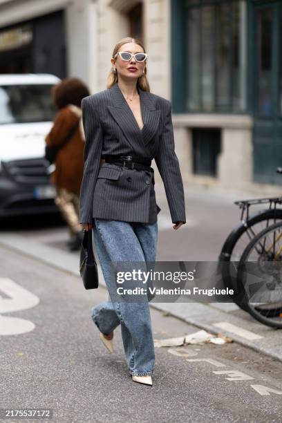 Guest wears white sunglasses, a black leather belt, a black leather bag, a grey striped blazer, blue straight jeans and white pumps outside Akris...