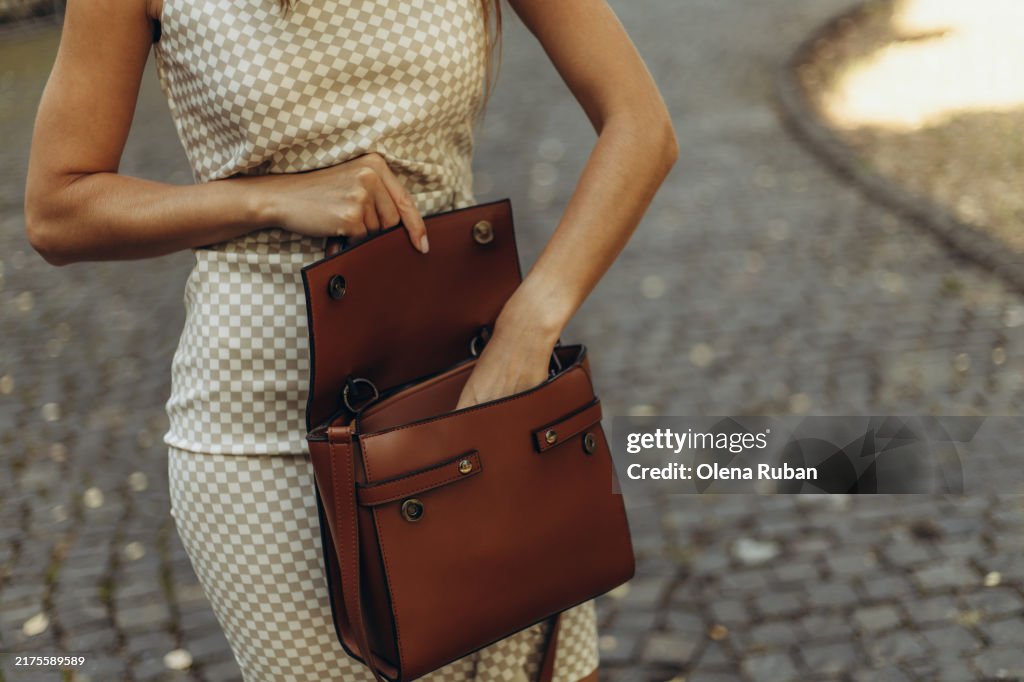 Young woman in a chess dress rummaging through a handbag on a stone walkway.