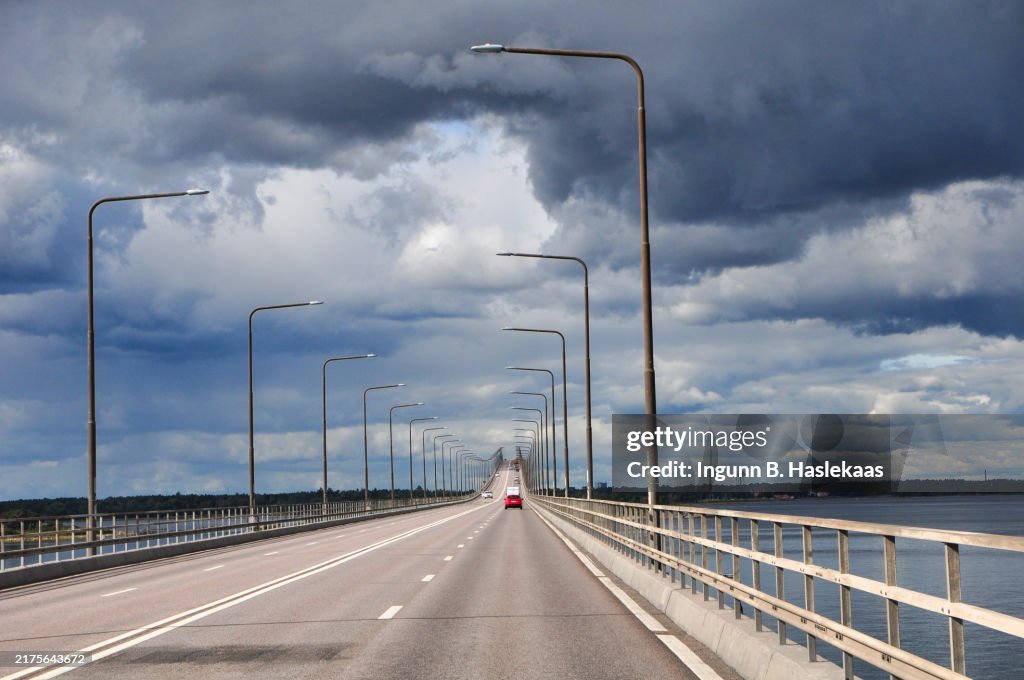 Red car and other vehicles on a very long bridge. Lamp lights. Cloudy sky. Multiple lanes and white line.
