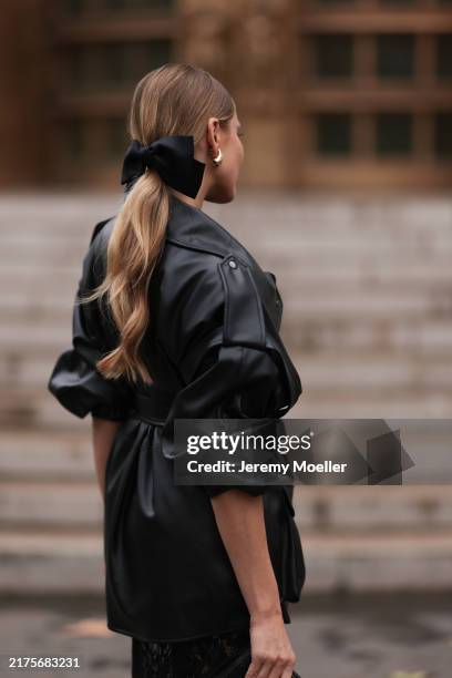 Ginta is seen wearing a black bow around her ponytail, gold drop-shaped earrings, a round diamond stud earring, an oversized black leather jacket...