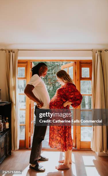 a sweet moment between an expectant couple - they compare baby bumps in a sunny room - nice abs stock pictures, royalty-free photos & images