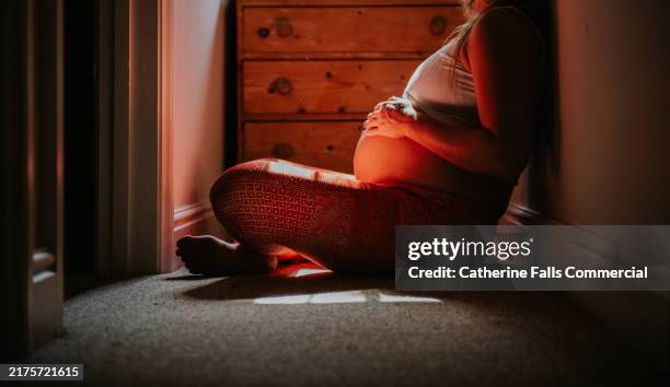 a pregnant woman sitting peacefully in a softly lit hallway, reflecting on motherhood at dusk - manos sobre el estómago fotografías e imágenes de stock