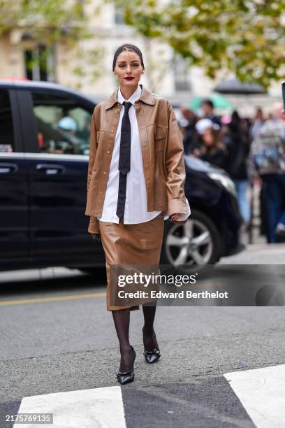 Olivia Palermo wears black headband gold earrings, white oversized buttoned up long sleeve shirt, black tie, shiny dark brown oversized Stella...