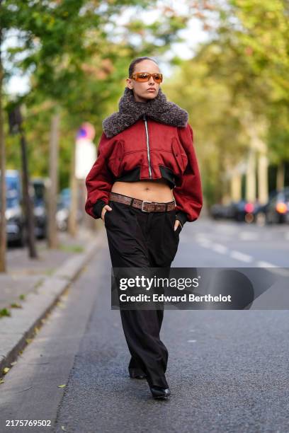 Guest wears orange sunglasses, dark red bomber jacket with dark brown fluffy collar, shiny dark brown leather belt, black loose pants, outside...
